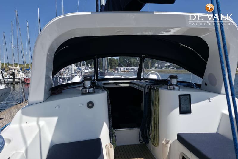 De foto toont het achterdek van een zeilboot met een blauwe buiskap, omringd door andere boten in een haven.
