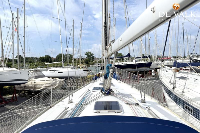 Een dek van een zeilboot met een mast, gieken, touwen, luiken en een blauwe afdekking, omringd door andere boten.
