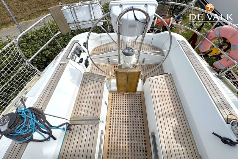 De foto toont de cockpit van een zeilboot met een stuurwiel, kompas, houten banken en een rooster op de vloer.