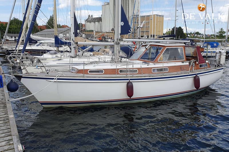 Een witte zeilboot met een houten kajuit en blauwe en rode strepen op de romp ligt aangemeerd in een haven. — Vilm 106