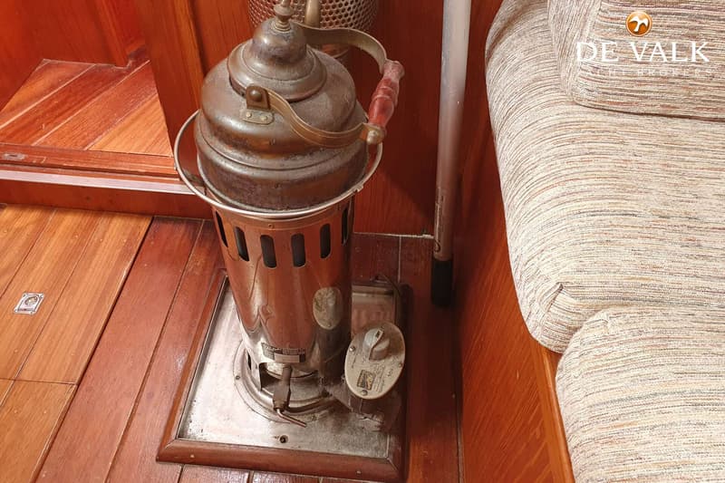Een koperkleurige kachel met een houten handvat staat op een houten vloer naast een gestreepte bank.
