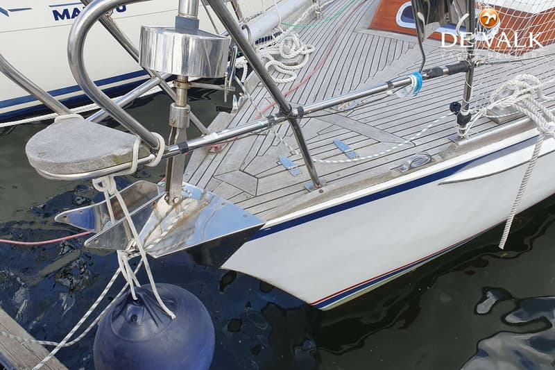 De foto toont de boeg van een witte zeilboot met een teakhouten dek, een ankerrol en een blauwe stootwil. — Vilm 106