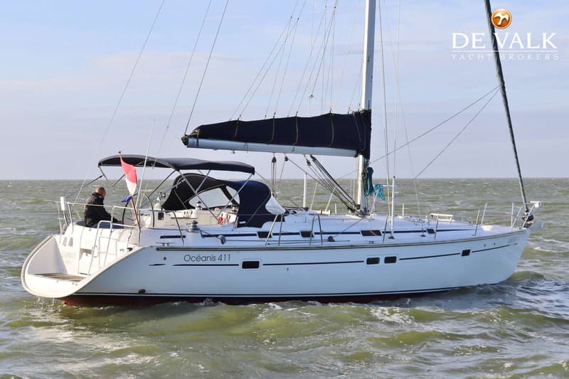 Een witte zeilboot met de naam "Océanis 411" vaart op water met een persoon aan het roer en een Nederlandse vlag.