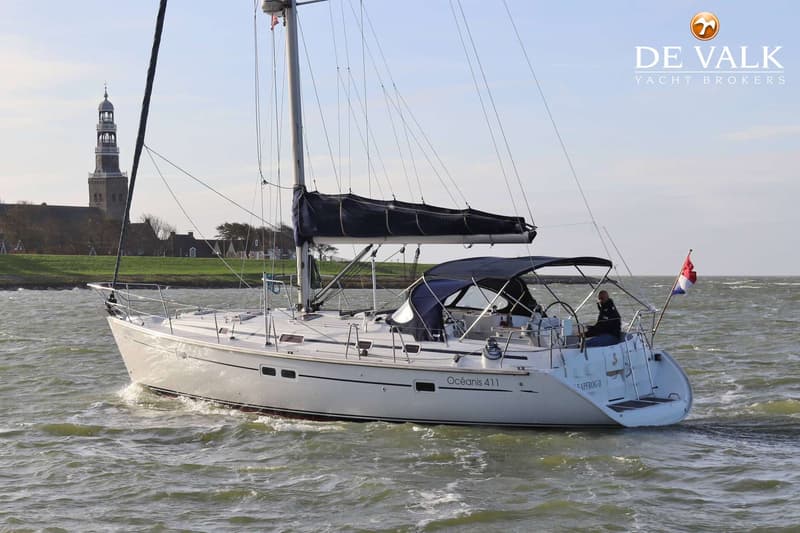 Een witte zeilboot met de naam "Océanis 411" vaart op water met een toren en landschap op de achtergrond.