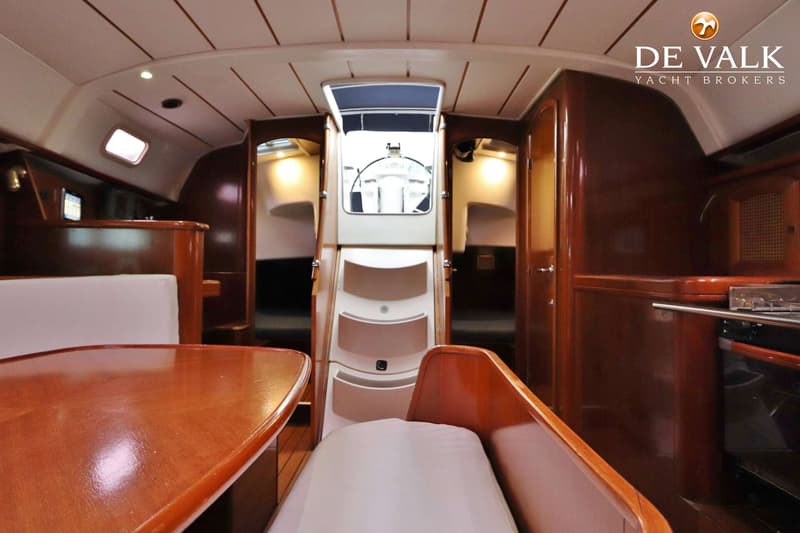 De foto toont het interieur van een boot met een houten tafel, banken, een trap naar de kuip en een kombuis.