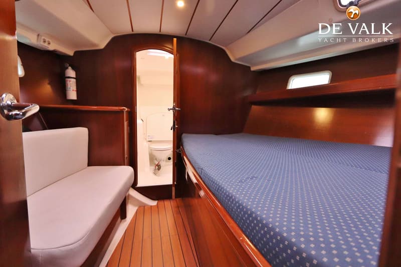 De foto toont een kajuit met een bed met blauw matras, een witte bank, een brandblusser en een open deur naar een…