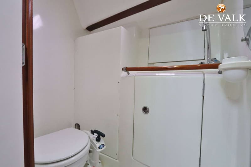 Een witte scheepstoiletruimte met een toilet, een wastafel met kraan en een houten handgreep.