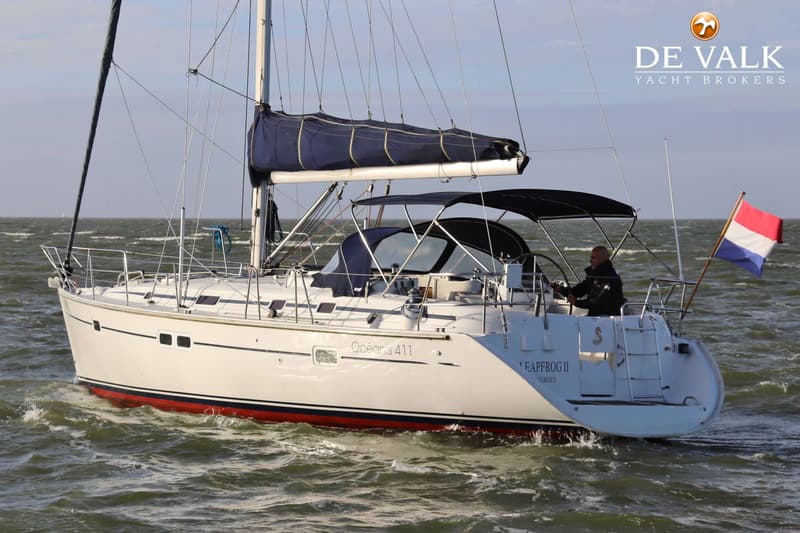 Een witte zeilboot met rode en blauwe strepen vaart op een golvende zee, met een man aan het roer en een Nederlandse vlag…