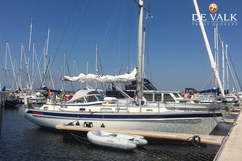 Een zeilboot met de naam "Flodebolle" ligt aangemeerd in een haven, met een bijboot ernaast. — Hallberg Rassy 39