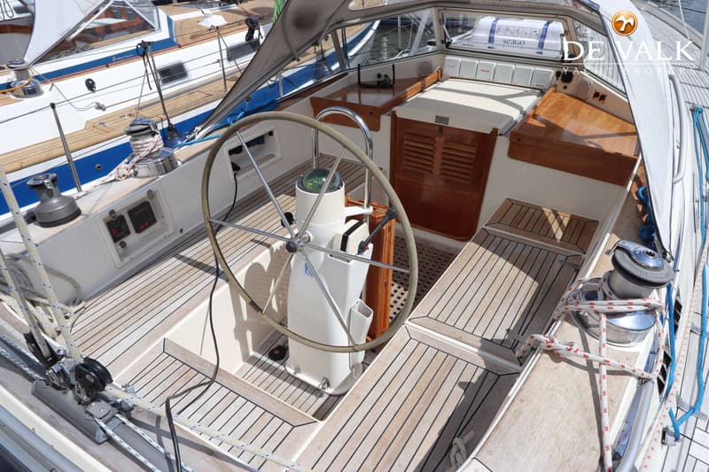 De foto toont de kuip van een zeilboot met een stuurwiel, kompas, teakhouten dek en banken, en diverse bedieningselementen.