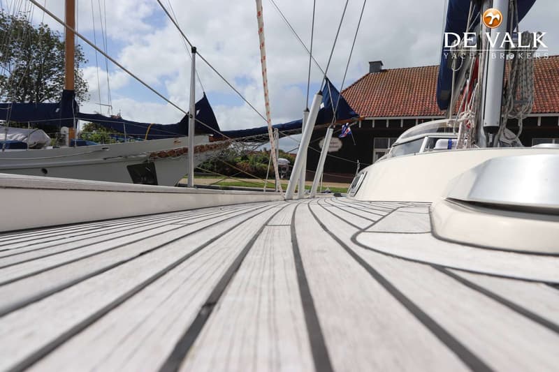 Een houten dek met zwarte naden, een mast en een kajuit zijn zichtbaar op een boot met een gebouw op de achtergrond.