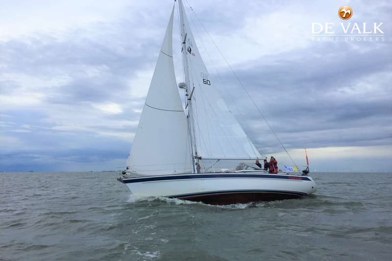 Een witte zeilboot met blauwe strepen en een bruine onderkant vaart op een grijze zee onder een bewolkte lucht.