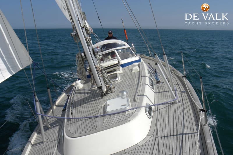 Een zeilboot met teakhouten dek vaart op zee, met een persoon aan het roer en een zeil deels zichtbaar. — Hallberg Rassy 39