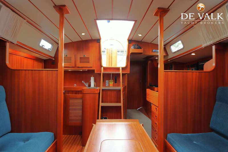 De foto toont het interieur van een boot met houten wanden, een tafel, blauwe kussens en een kombuis. — Hallberg Rassy 39