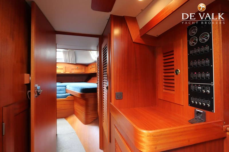 Een interieur van een boot met houten wanden, een open deur naar een slaapcabine met blauwe kussens en een schakelpaneel.