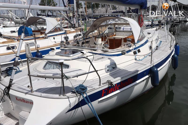 Een witte zeilboot met blauwe strepen en de naam "Flødebolle" in rode letters ligt in het water. — Hallberg Rassy 39