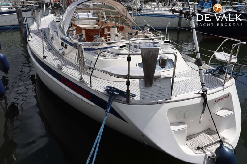 Een witte zeilboot met een teakdek en een blauwe streep langs de romp ligt aangemeerd in het water. — Hallberg Rassy 39