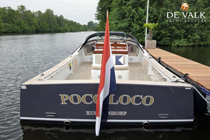 Een boot met de naam "POCO LOCO" en "KORINHOEF" op de spiegel, met een Nederlandse vlag, ligt afgemeerd aan een houten…