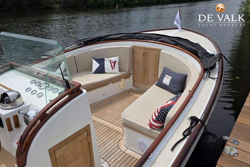 Een bovenaanzicht van een boot met een houten dek, beige kussens en een stuurconsole, liggend aan een houten steiger.