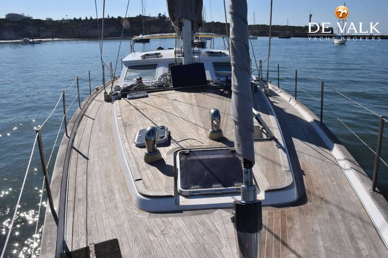 Een houten dek van een zeilboot met een mast, dekramen en reling is zichtbaar, met water en land op de achtergrond. — CRN 63