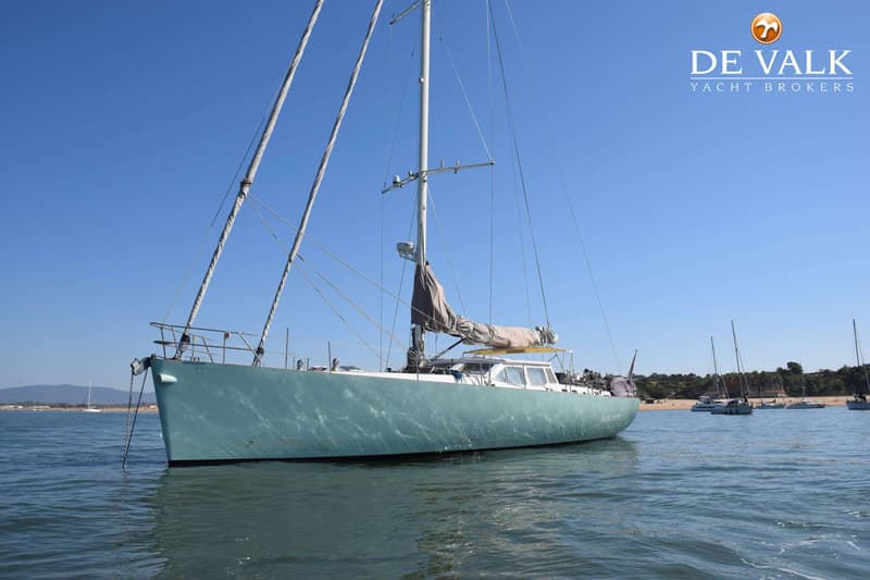 Een lichtgroene zeilboot met opgerolde zeilen en een ankerketting hangt in helder water onder een blauwe lucht, met andere…