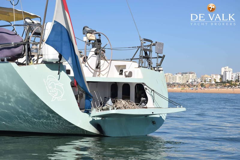 De achtersteven van een lichtblauwe zeilboot met een Nederlandse vlag, stuurwiel en de naam "mahi mahi" op de romp is…