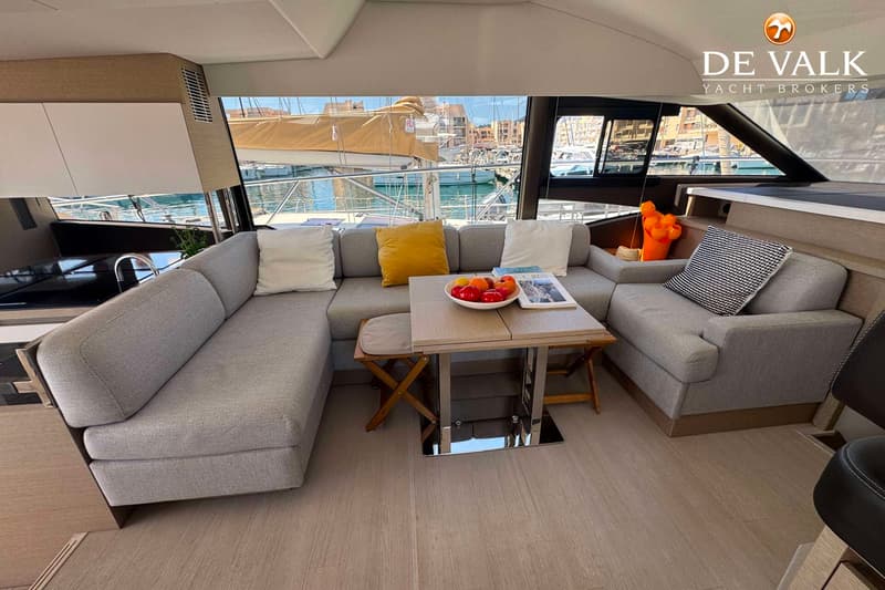Een interieur van een boot met een grijze hoekbank, een salontafel met fruit en tijdschriften, en uitzicht op een haven.