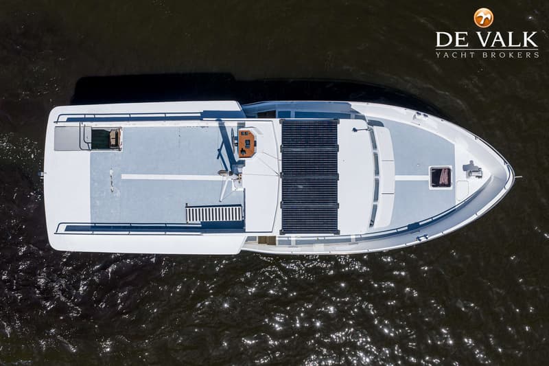 Een bovenaanzicht van een witte boot met een lichtblauw dek, zonnepanelen en een stuurstand, omringd door donker water.