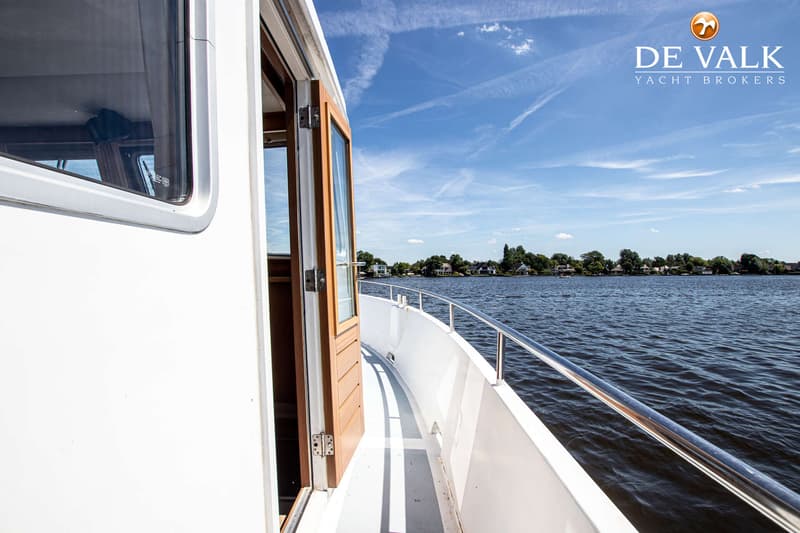 Een open houten deur leidt naar het interieur van een witte boot met een reling, varend op water met bebouwde oevers.