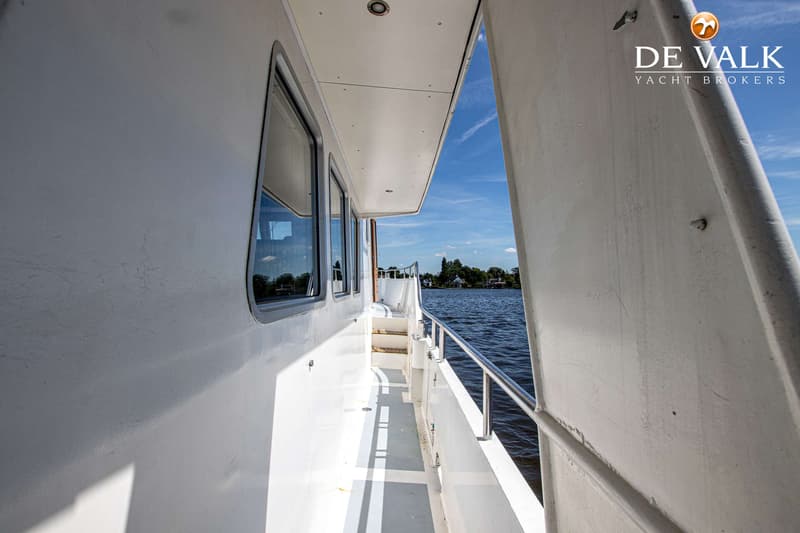 Een zijaanzicht van een boot met ramen, een reling en een trap, varend op een waterweg met bebouwing op de achtergrond.