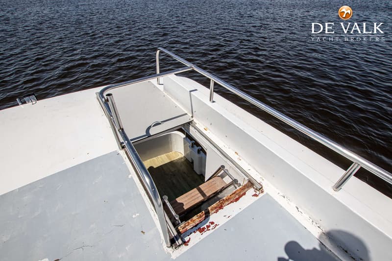 Een open luik op het dek van een boot, met een houten bankje en een metalen reling, en water op de achtergrond.