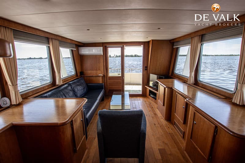 Een interieur van een boot met houten vloer en wanden, een donkerblauwe bank, een fauteuil, een glazen salontafel en ramen…