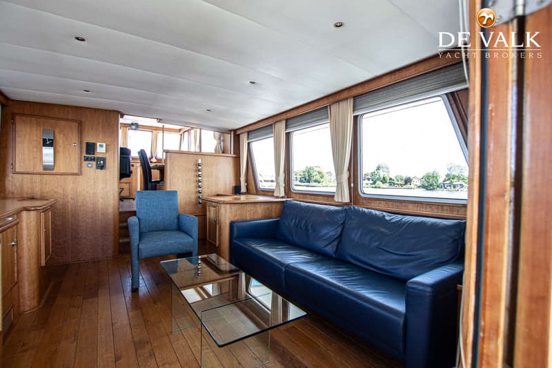 De foto toont het interieur van een boot met houten wanden en vloer, een blauwe bank, een blauwe stoel en een glazen…