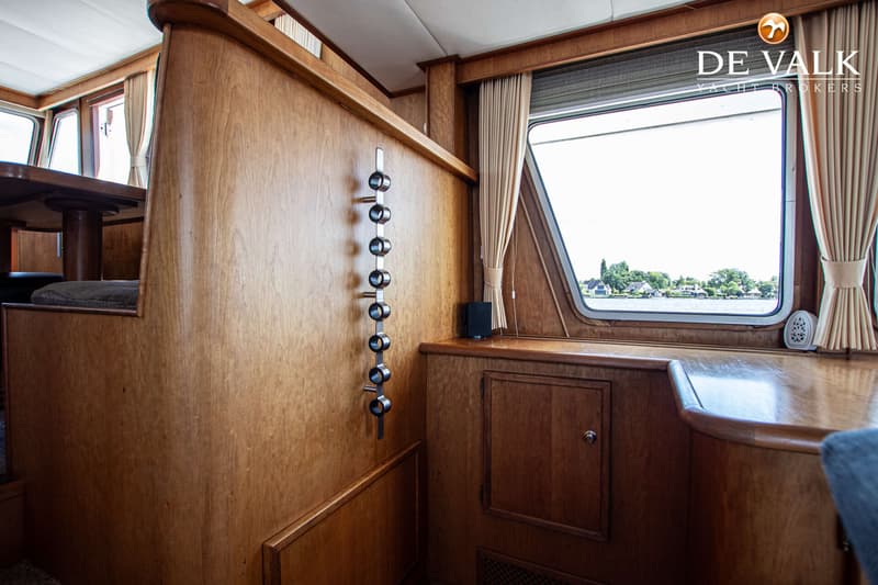 De foto toont het interieur van een boot met houten wanden, een raam met uitzicht op water en huizen, en een rek met zes…