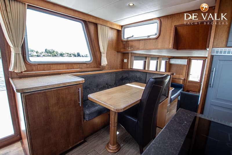 Een interieur van een boot met houten wanden, een tafel, een bank en een stoel, en ramen met uitzicht op water en huizen.