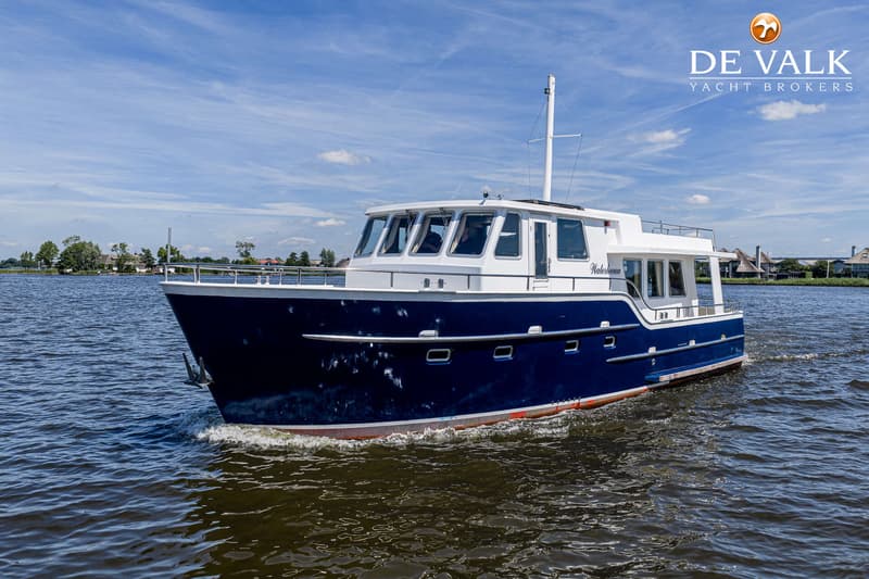 Een blauw-witte motorboot met de naam "Waterlelie" vaart op een waterweg onder een blauwe lucht met wolken.