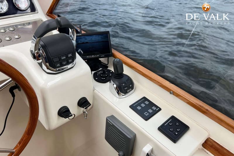 De foto toont een deel van een bootstuurhut met een stuurwiel, bedieningspanelen, een monitor en uitzicht op water.