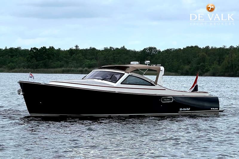 Een donkerblauwe boot met een witte opbouw en een beige cabriokap vaart op water met een bosrijke oever op de achtergrond.