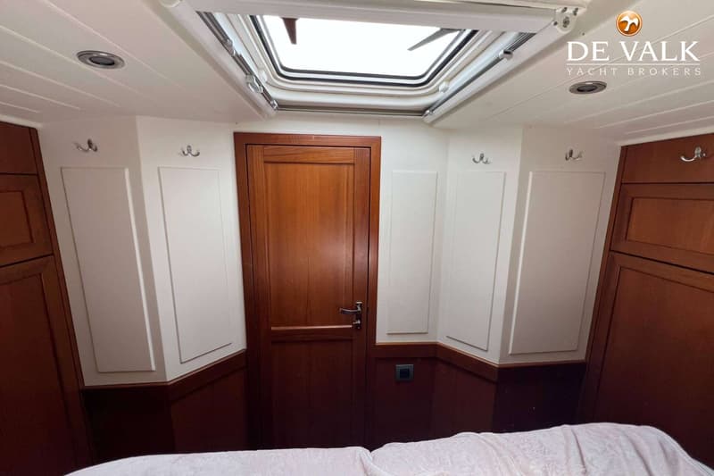 Een kajuit met een houten deur, een dakluik, witte wanden met houten accenten en een bed met een wit dekbed.