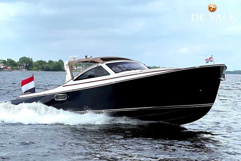 Een donkerblauwe motorboot met witte opbouw en bruine kap vaart over water, met een Nederlandse vlag aan de achtersteven.