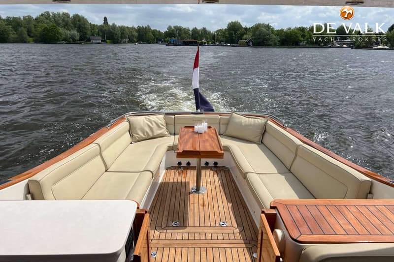 Een boot met een houten dek en beige kussens vaart over water, met een Nederlandse vlag aan de achterkant.
