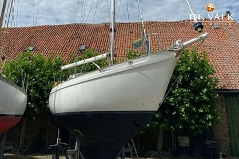 Een witte zeilboot met een donkere romp staat op bokken voor een gebouw met een pannendak en struiken. — Tradewind 35