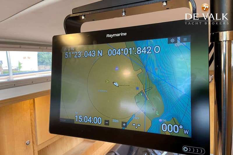 Een Raymarine plotter toont een kaart met coördinaten, tijd, diepte en koers, gemonteerd in een houten interieur.