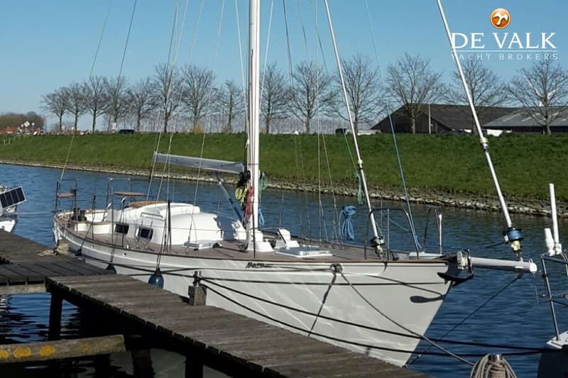 Een witte zeilboot ligt afgemeerd aan een houten steiger, met een groen talud en bomen op de achtergrond. — Tradewind 35