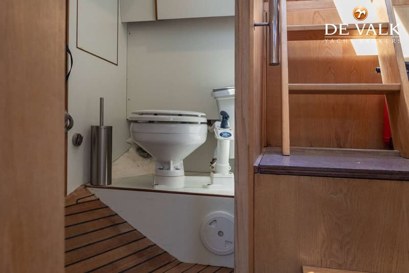 Een witte boottoilet met een Jabsco-pomp en een toiletborstel staan op een houten vloer naast een houten trap.