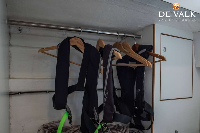 Drie zwarte reddingsvesten hangen aan houten hangers aan een metalen stang, met touwen eronder en een witte deur rechts.
