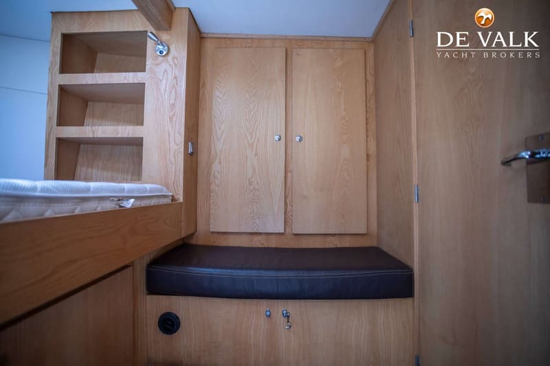 De foto toont een kajuitinterieur met houten wanden, een bed met matras, open schappen, een bankje en kasten.