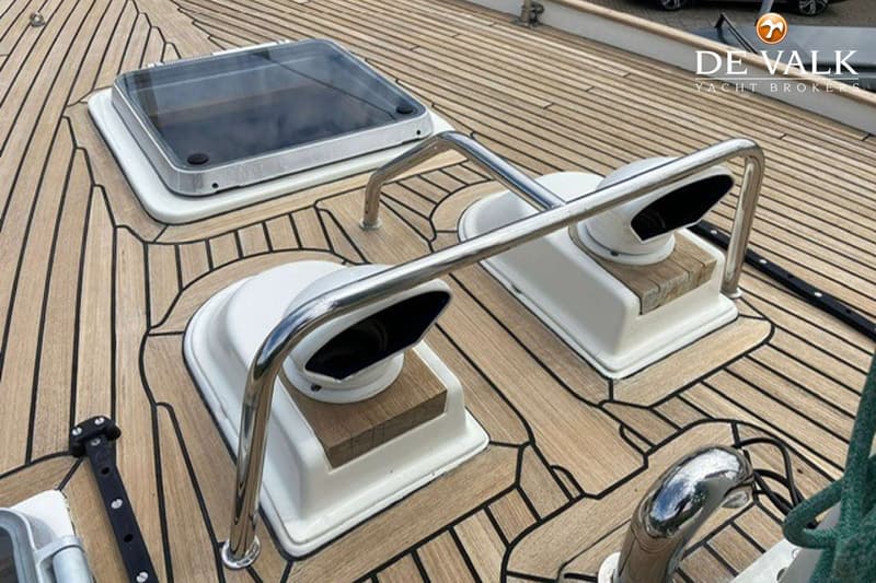 Een houten dek met zwarte naden, een luik, twee ventilatiekappen en een RVS beugel zijn zichtbaar.