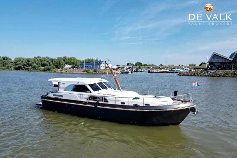 Een donkerblauwe motorboot met witte opbouw vaart op een waterweg, met een jachthaven en gebouwen op de achtergrond.