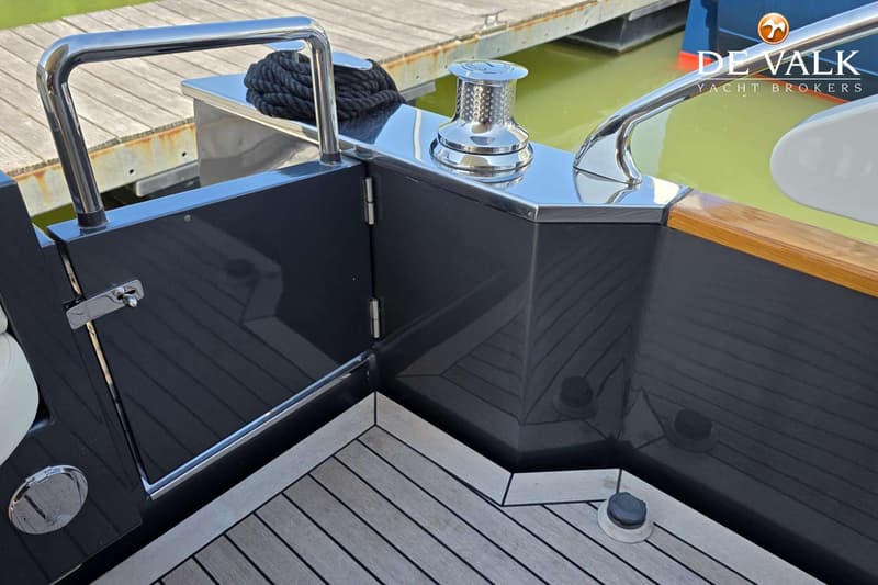 Een detailfoto van een boot met een teakhouten dek, een glanzende grijze romp, een lier en een bolders.